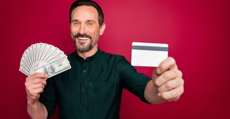 Close-up portrait of his he nice cheery bearded man holding in hand giving you freelance plastic card withdrawal isolated on bright vivid shine vibrant maroon burgundy marsala red color background