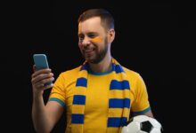 Studio portrait of smiling lucky football fan man holding ball in his hand and making bets on favorite team at bookmaker's website using smartphone. Isolated over blacl background.
