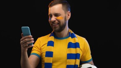 Studio portrait of smiling lucky football fan man holding ball in his hand and making bets on favorite team at bookmaker's website using smartphone. Isolated over blacl background.